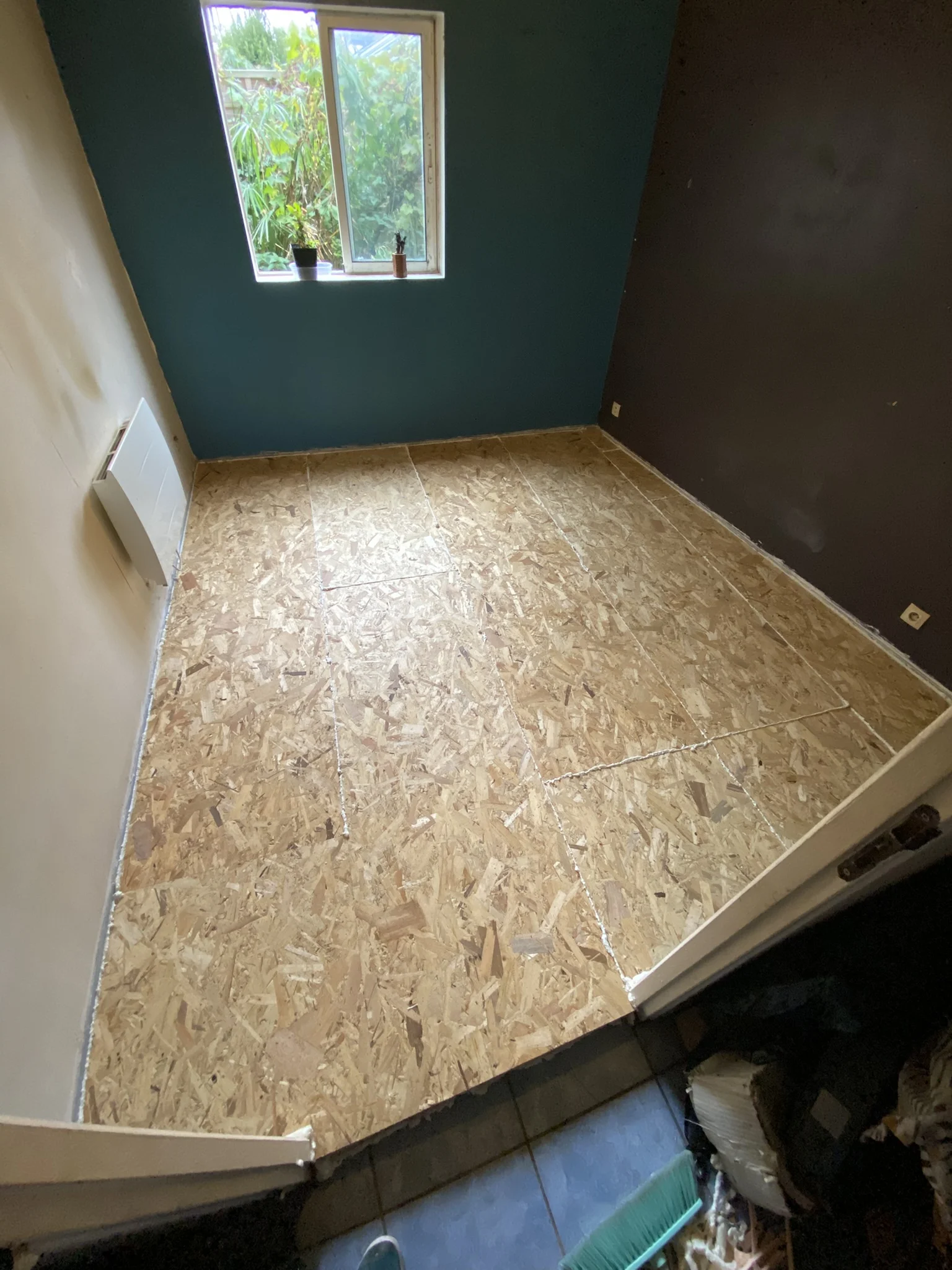 Chambre vide avec sol recouvert de panneaux de bois OSB, murs peints en bleu, beige et gris, fenêtre avec vue sur des plantes.