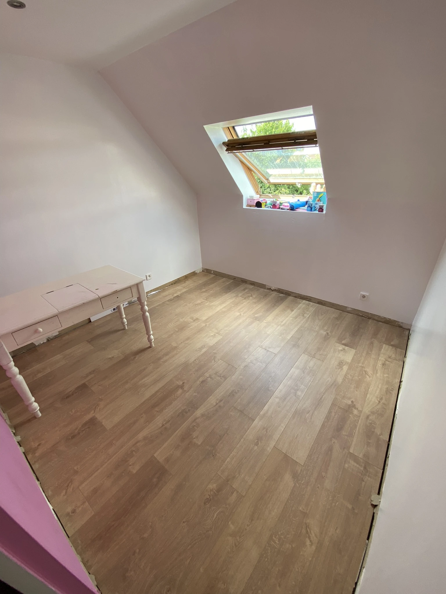 Chambre vide avec parquet en bois clair, une fenêtre de toit ouverte et un petit bureau blanc contre un mur blanc
