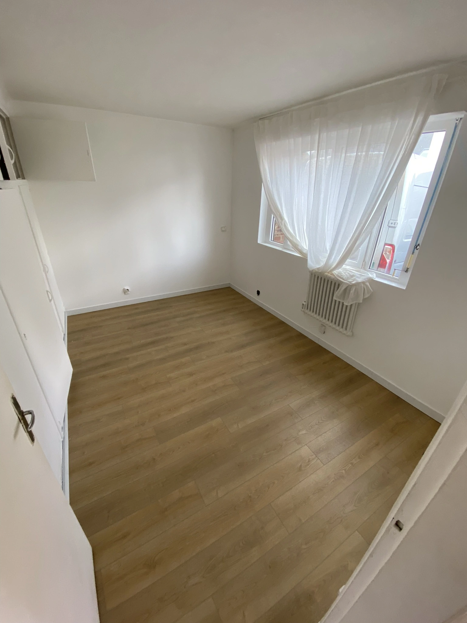 Chambre vide avec sol en bois clair, murs blancs, fenêtre ouverte avec rideau blanc et radiateur sous la fenêtre