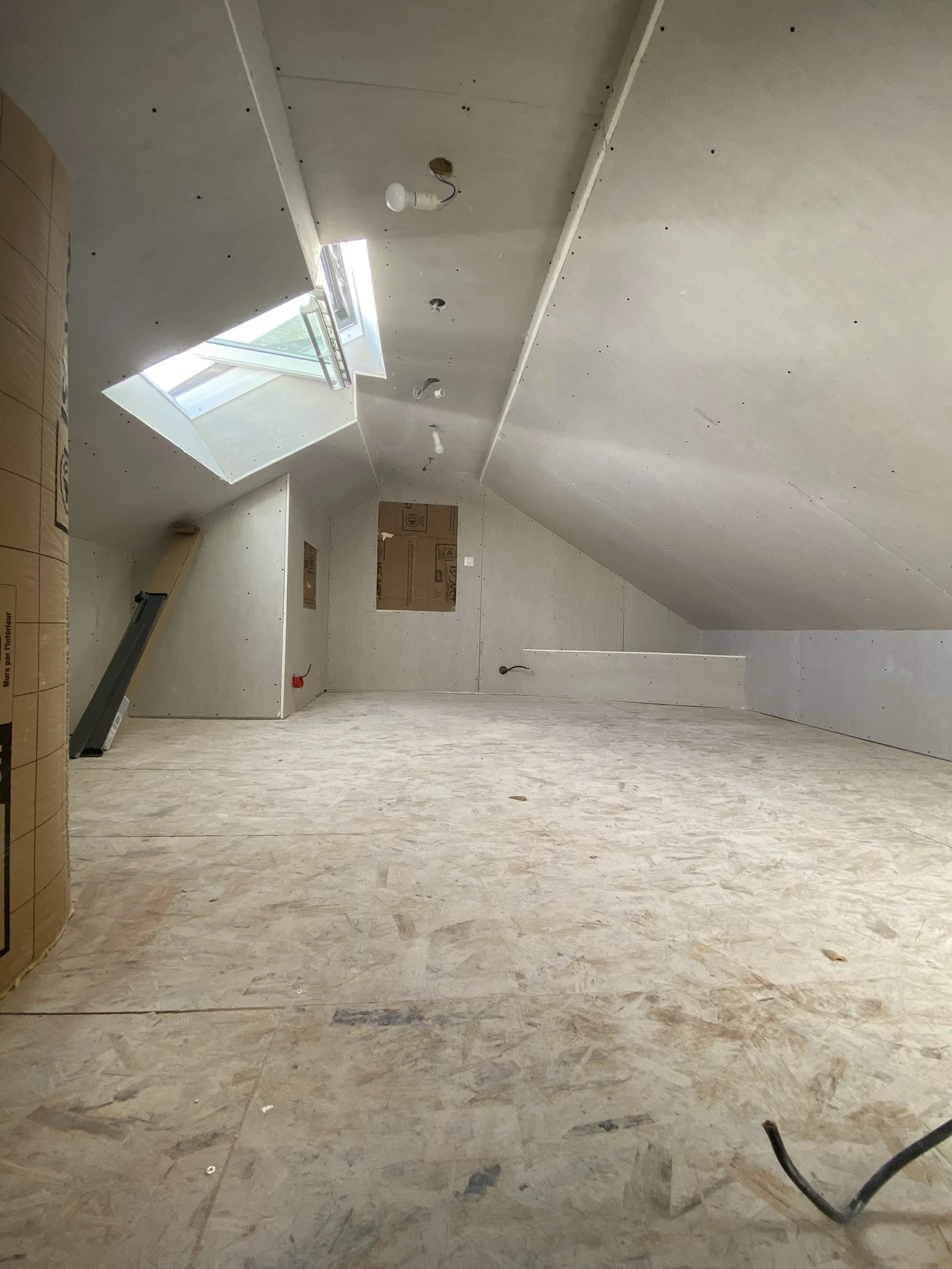 Combles non aménagés avec plancher en bois brut, murs et plafond en plaques de plâtre, fenêtre de toit ouverte.