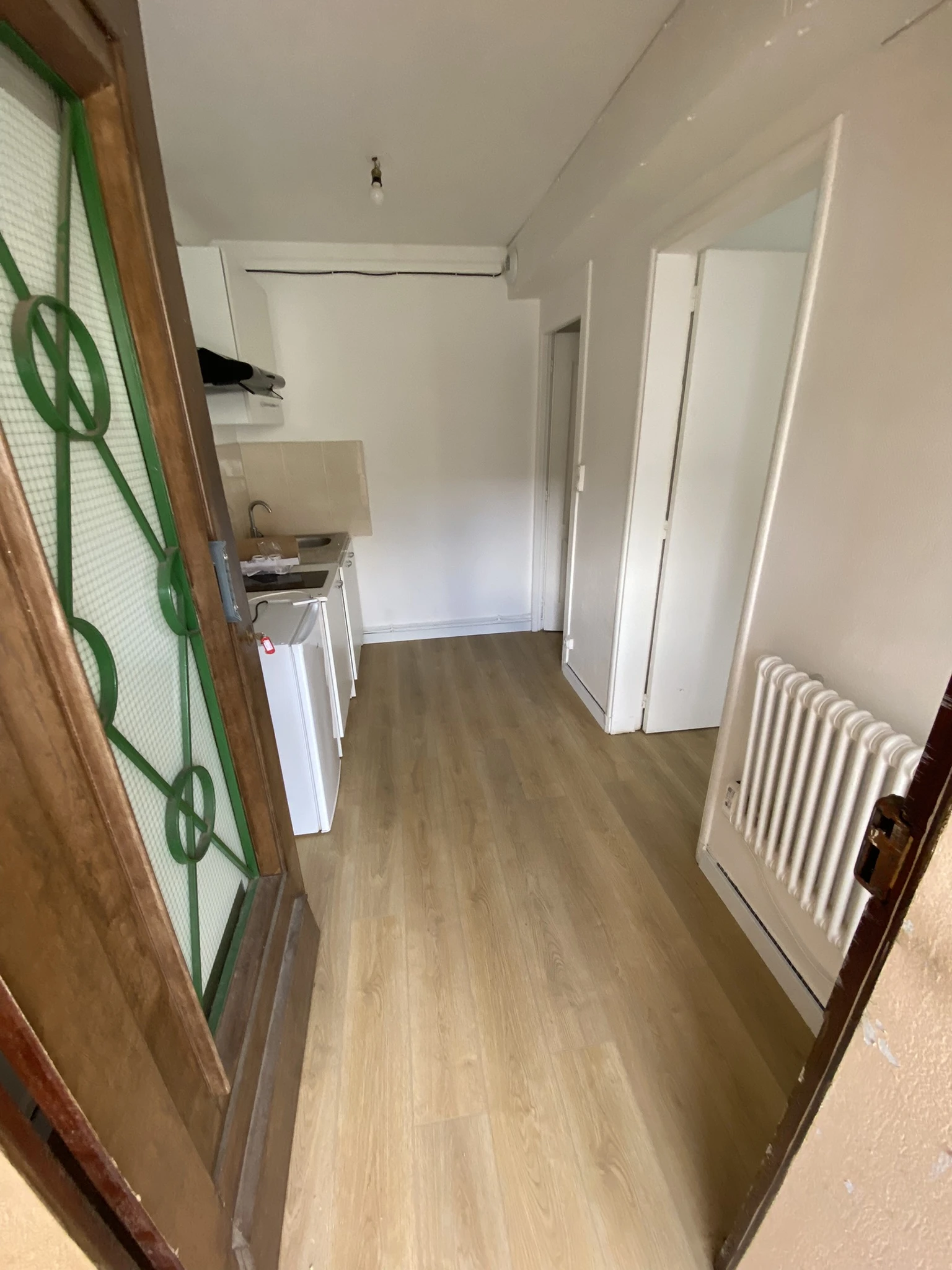 Vue d'un couloir avec un sol en bois clair, une cuisine équipée d'un évier et d'un petit réfrigérateur à gauche, et un radiateur blanc à droite.