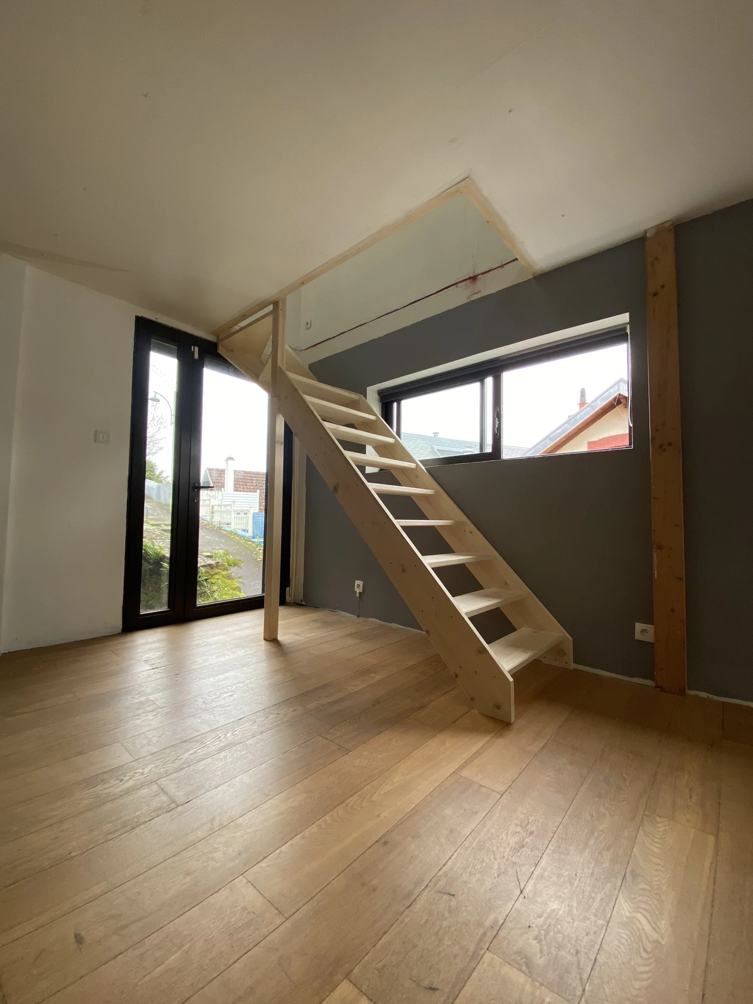 Escalier en bois clair menant à une mezzanine dans une pièce avec sol en parquet, mur gris et grande fenêtre.
