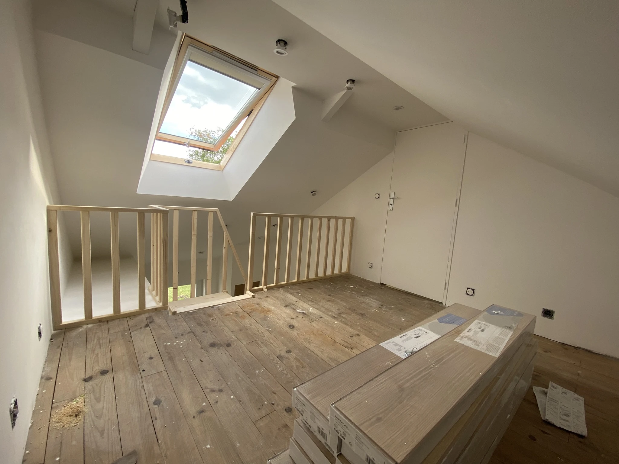 Espace sous combles avec plancher en bois, rambarde en bois clair, fenêtre de toit ouverte et murs blancs en cours de rénovation