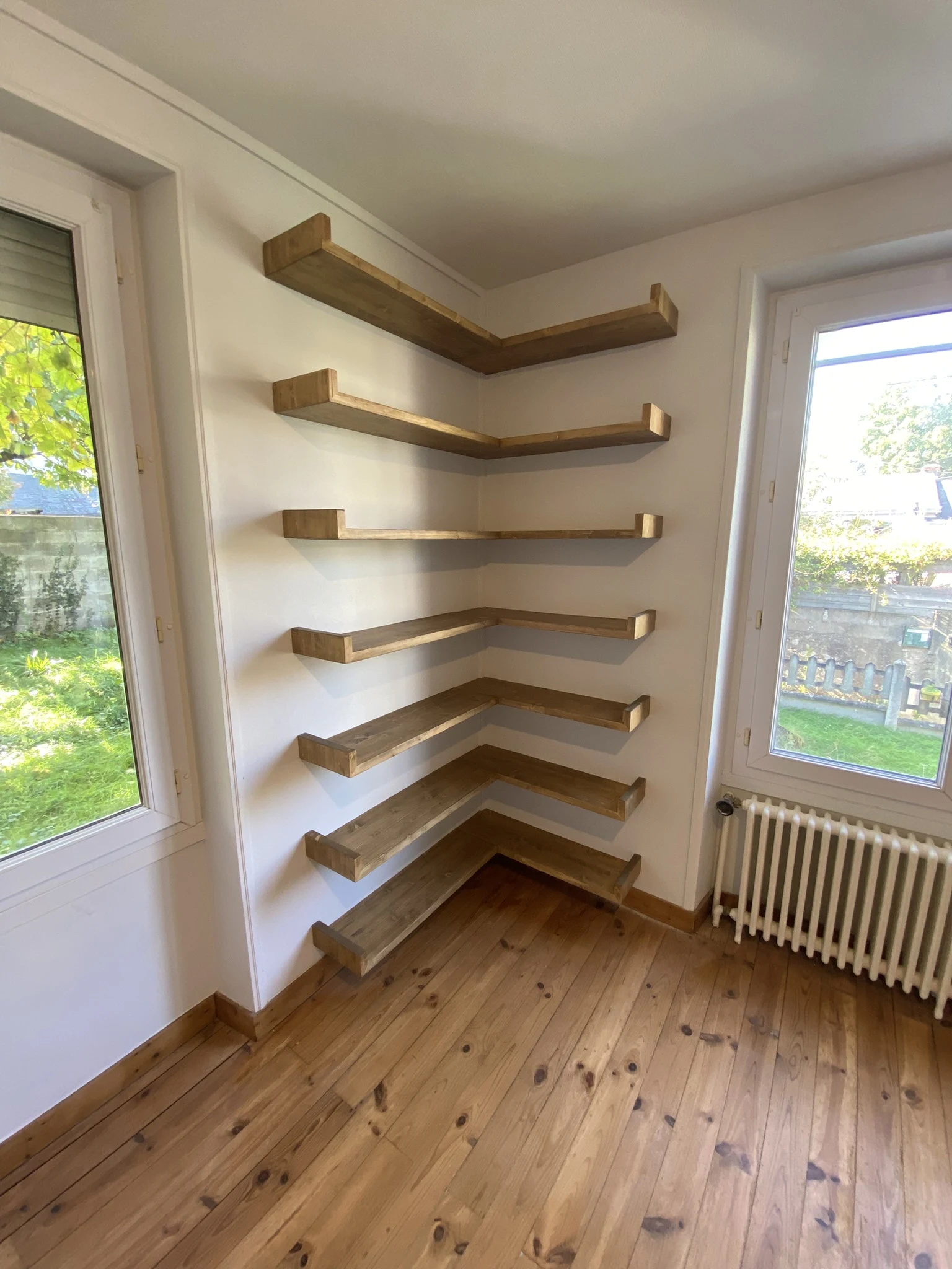 Étagère d'angle en bois fixée sur un mur blanc dans une pièce avec parquet et fenêtres donnant sur un jardin