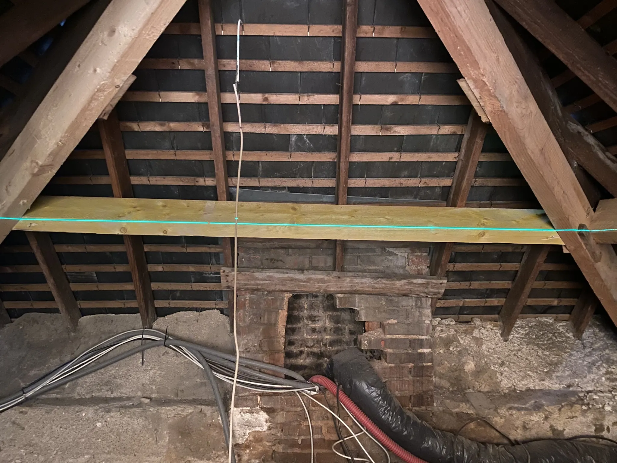 Intérieur d'un grenier avec charpente en bois, mur en briques partiellement endommagé et plusieurs câbles électriques visibles