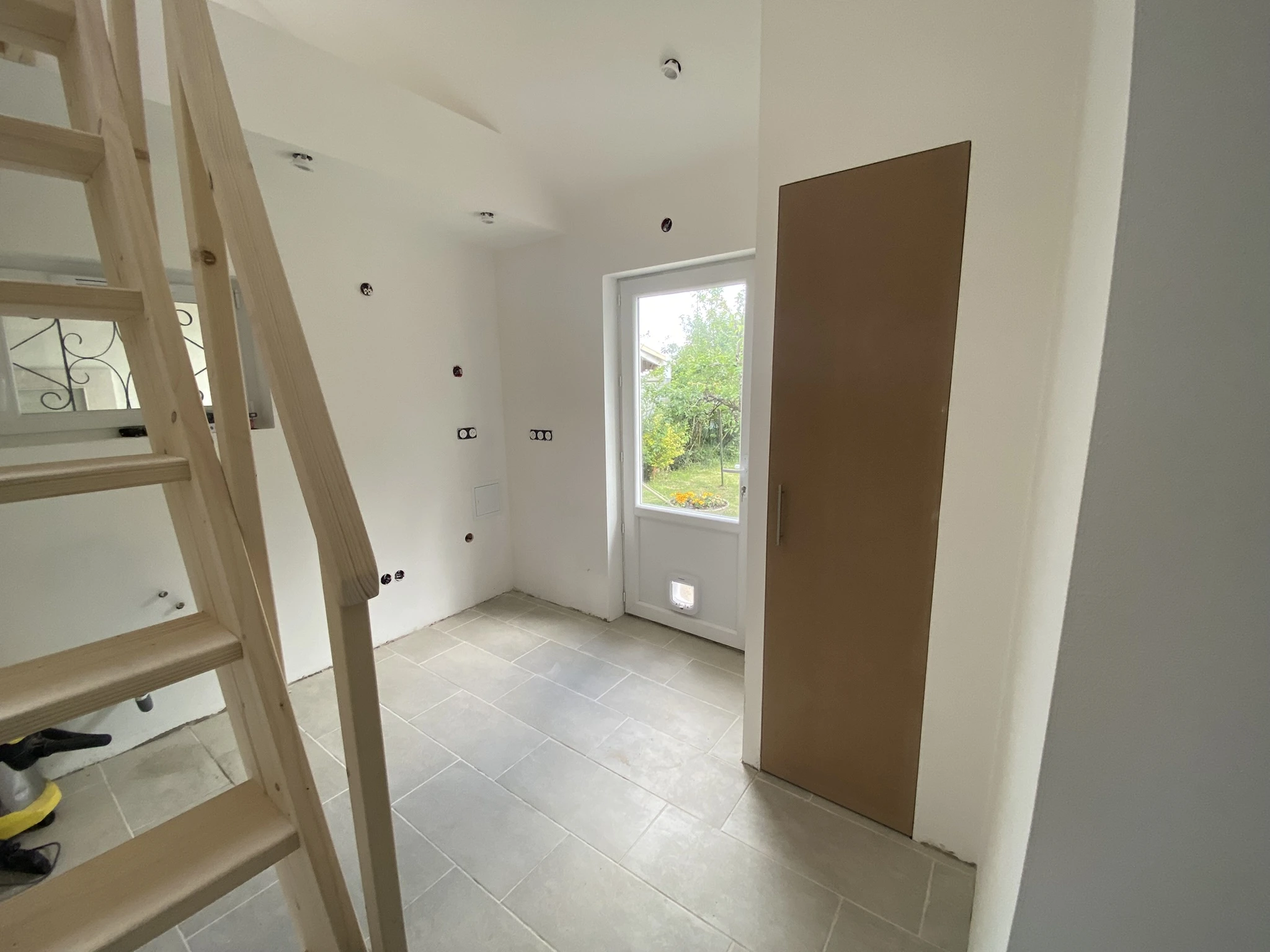 Pièce vide avec escalier en bois à gauche, porte-fenêtre donnant sur un jardin, sol carrelé gris et murs blancs.